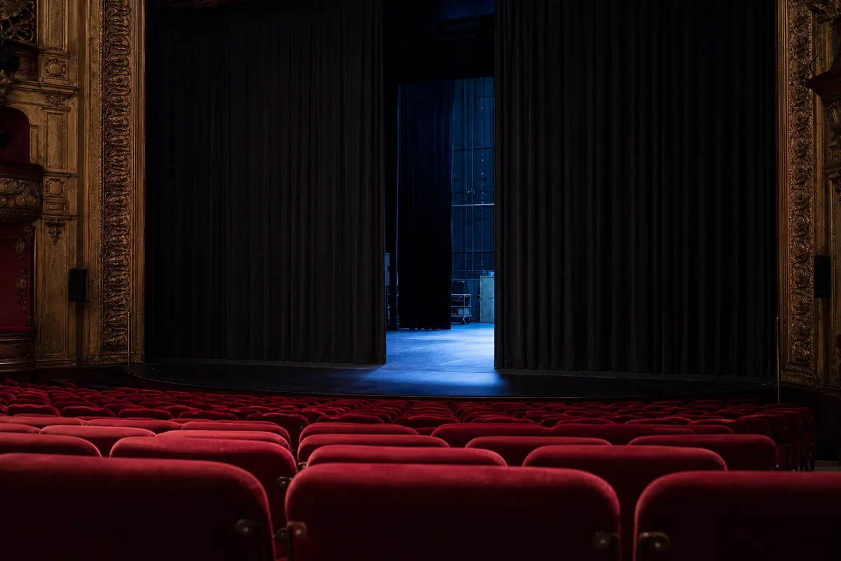 Plateau de théâtre nu vu depuis la salle, rideau sombre entrouvert et lumière de service